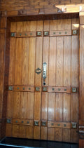 A pair of large, traditional wooden double doors with a warm medium-brown finish, featuring decorative brass hardware, metal studs, and a central metal latch.